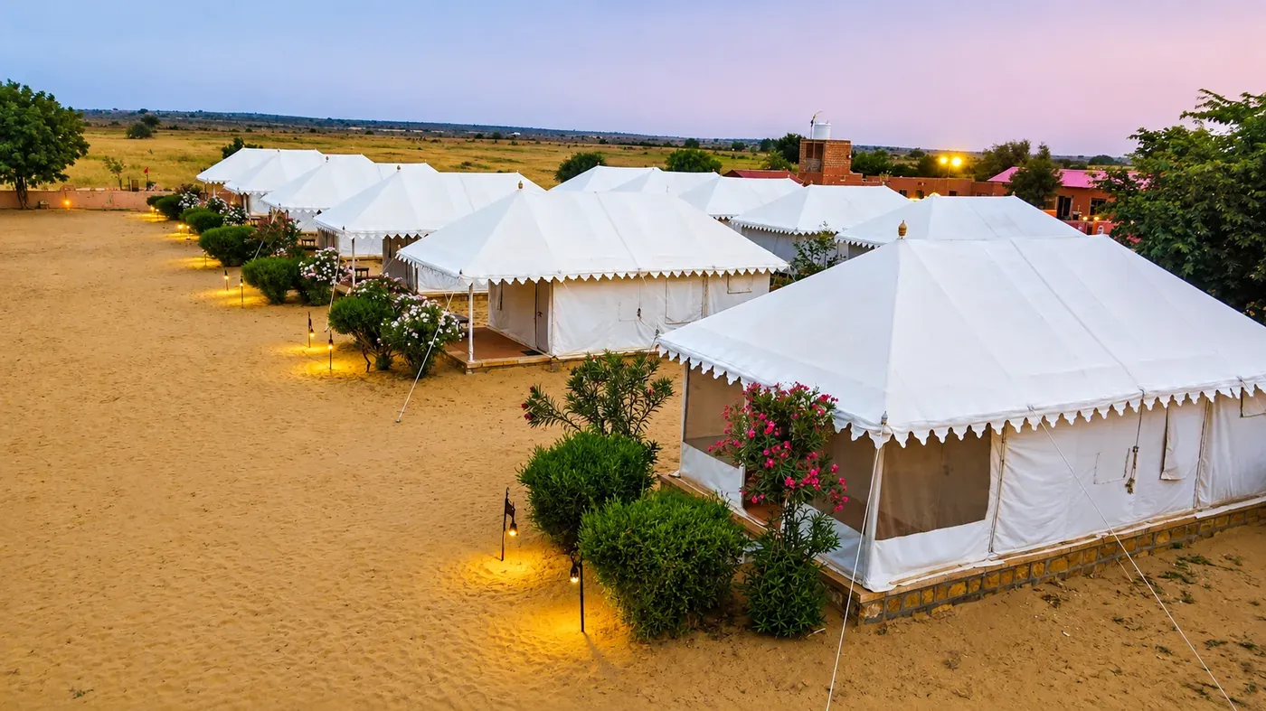 White Swiss glamping tents in desert sand grounds with garden shrubs at sunset